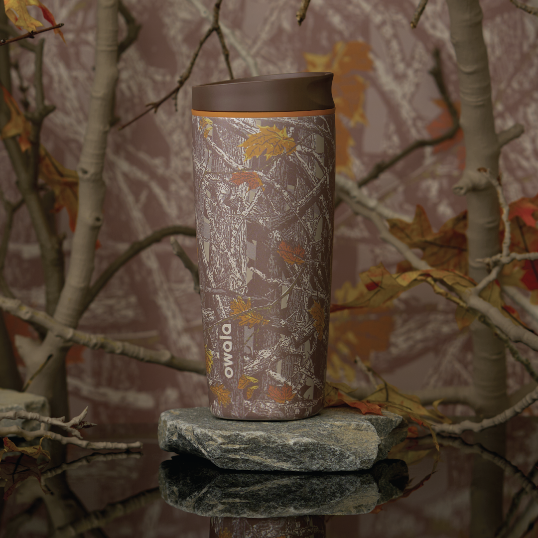 SmoothSip Slider in Out of Sight, woodland camo patterned body in brown and tan with matching brown flip cap and carry loop, shown on a mossy rock against a forest backdrop